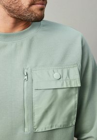 Sudadera verde menta con un tejido texturizado, que cuenta con un bolsillo con cremallera y un gran bolsillo con botón en el pecho.