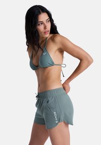 Haut de bikini vert à bonnets triangulaires ; short de bain assorti vert avec cordon de serrage et poche latérale ; détail du logo sur le short.