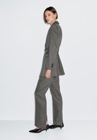 Szary, dopasowany garnitur z długą marynarką i prostymi spodniami. Gładki materiał i wyrafinowany krój, uzupełniony czarnymi szpilkami.