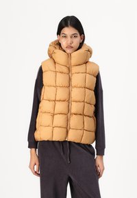 Gilet senza maniche in un colore marrone dorato, dotato di cappuccio, design trapuntato e zip frontale. Indossato sopra un abbigliamento scuro a coste.