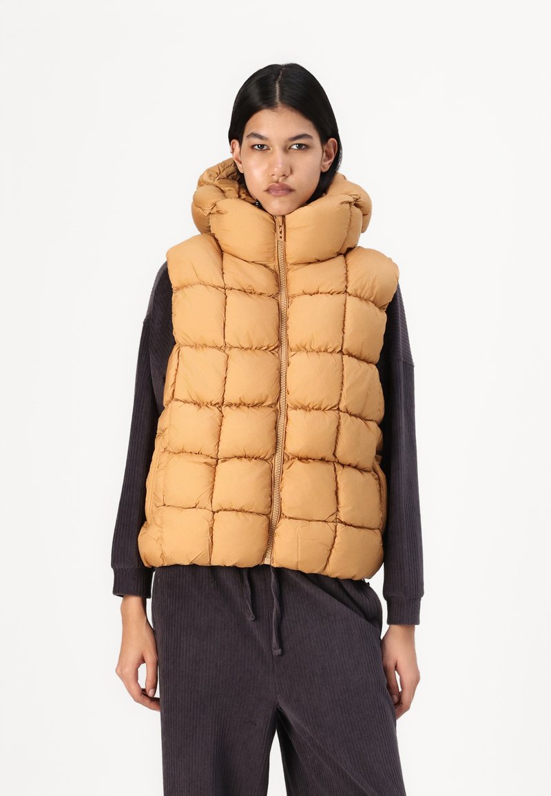 Gilet senza maniche in un colore marrone dorato, dotato di cappuccio, design trapuntato e zip frontale. Indossato sopra un abbigliamento scuro a coste.