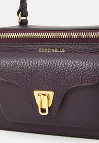 Purple leather handbag featuring a textured surface, gold zip closure, and a gold accent lock; branded with "Coccinelle" on the front.