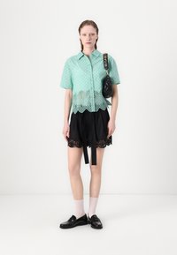 Korte mouw mintgroene kant blouse met een golvende zoom, gecombineerd met een zwarte korte broek met kantafwerking en een stoffen riem. Zwarte accessoire tas.