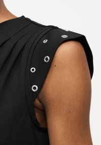 Μπλούζα χωρίς μανίκια σε μαύρο χρώμα με ασημί μεταλλικά περικάρπια κατά μήκος της ραφής του ώμου σε ένα χέρι και ώμο ατόμου που φαίνεται πάνω σε λευκό φόντο.