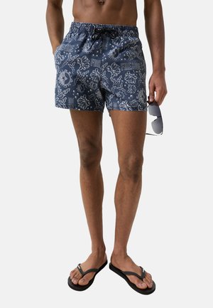 Mann trägt marineblaue gemusterte Badeshorts, schwarze Flip-Flops, hält schwarze Sonnenbrille mit reflektierenden Gläsern in der rechten Hand.