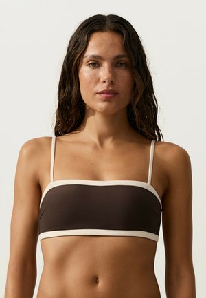 Kvinde med langt mørkt bølget hår iført en brun bandeau-top med hvid kant, vendt fremad mod en ensartet lys baggrund.