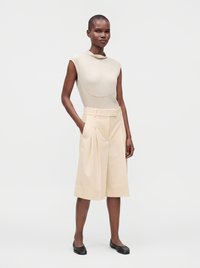 Femme debout portant un haut beige sans manches et un short large beige clair jusqu'aux genoux avec des ballerines noires sur fond blanc.