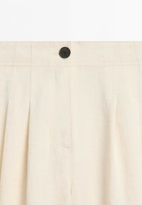 Close-up of cream-colored pants with pleats, stitched waistband, and a single dark button at the center front.
