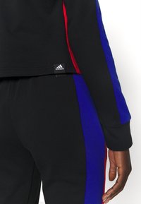 Pantalon de sport noir et haut avec des rayures verticales bleues et rouges, montrant une étiquette de logo Adidas sur l'ourlet du t-shirt près de la taille.