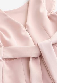 Tissu rose doux avec une fermeture éclair latérale cachée et une large ceinture nouée en nœud sur un vêtement à texture lisse.