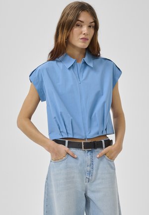 Kobieta w bezrękawniku jasnoniebieskiej, krótkiej, zapinanej na zamek koszuli z kołnierzykiem, w połączeniu z jasnoniebieskimi, wysokim stanem dżinsami i czarnym paskiem, z rękami w kieszeniach.