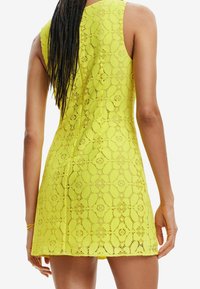 Vestido de encaje amarillo con patrones florales, diseño sin mangas y una silueta ajustada. La textura es intrincada, con una tela ligera y transpirable.