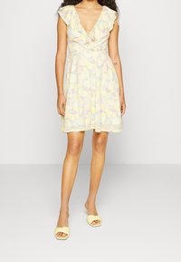 Robe jaune pastel à motif floral avec manches à volants et col en V profond. Silhouette évasée, associée à des sandales à enfiler jaune clair.