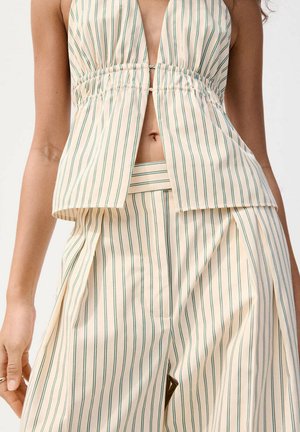 Femme portant une tenue crème deux pièces avec des rayures verticales vertes, comprenant un haut à dos nu avec découpe et un pantalon large assorti.