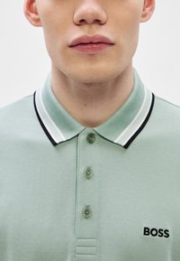 Polo de cor verde pálido com tecido texturizado, gola preta e branca, três botões e logótipo "BOSS" bordado a preto.