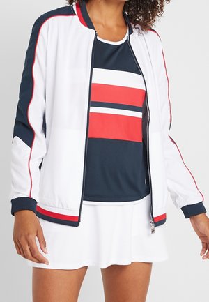 Vrouw draagt een wit tennisjack met rode en marineblauwe strepen over een marineblauw, rood en wit gestreept topje, gecombineerd met een witte tennisrok.
