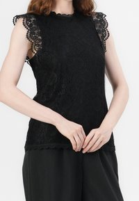 Svart spets ärmlös topp med scallopkanter, blommönster och texturerade detaljer, i kombination med lösa svarta byxor.