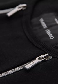 Primer plano de una prenda negra con cremallera, que presenta un herraje de cremallera plateado y una textura de tela suave, con la etiqueta de la marca visible en el interior.