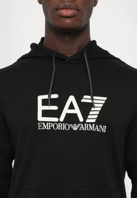 Felpa nera con logo ricamato bianco "EA7 EMPORIO ARMANI". Presenta cappuccio con coulisse e tessuto testurizzato. Design casual classico.