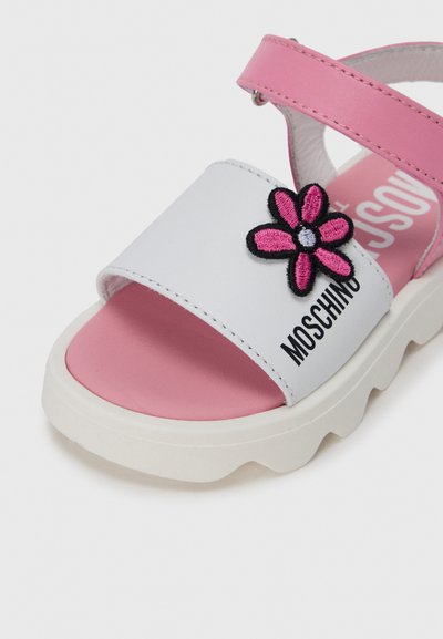 Λευκό και ροζ σανδάλι με λεπτομέρεια κεντήματος με λουλούδια, ανυψωμένη σόλα και ρυθμιζόμενο λο υράκι· διαθέτει λογότυπο στο λουράκι και υφές υλικών.
