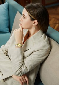 Reloj de pulsera dorado con una esfera redonda, banda articulada; traje beige claro; anillo dorado sencillo; pendientes colgantes con un acento azul claro.