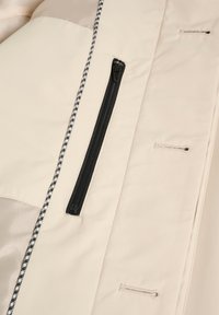 Abrigo beige con un bolsillo con cremallera negro, tejido texturizado, costuras destacadas y acentos en drawstring de cuadros en contraste.