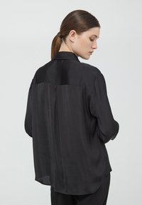 Camicia di seta nera a maniche lunghe con colletto su misura, vestibilità leggermente ampia e un pannello in contrasto nella parte superiore della schiena. Texture morbida.