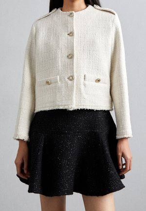 Woman wearing a white textured buttoned jacket with gold buttons and a black flared skirt on a gray background.