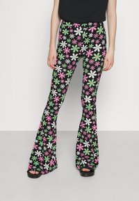 Vrouw in zwarte flared broeken met een groen, roze en witte bloemenprint, gecombineerd met zwarte open-teenschoenen en een zwarte mouwloze top.