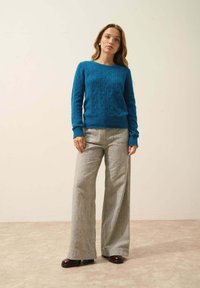 Turkos stickad tröja med ett texturerat diamants mönster, kombinerad med ljusgrå vida byxor och mörkbruna platåskor.