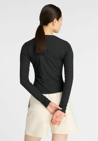 Schwarz langärmliges Oberteil mit taillierter Passform und strukturierter Stoffqualität. Kombiniert mit hochgeschnittenen beigen Shorts, die eine elegante Rückenansicht präsentieren.