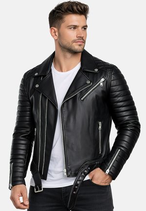 Un hombre con una chaqueta motera de cuero negro y una camiseta blanca está de pie con una mano en el bolsillo y mira hacia un lado sobre un fondo sencillo.