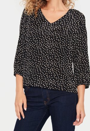 Mujer con blusa negra de escote en V con lunares irregulares blancos y mangas tres cuartos, combinada con vaqueros azul oscuro.