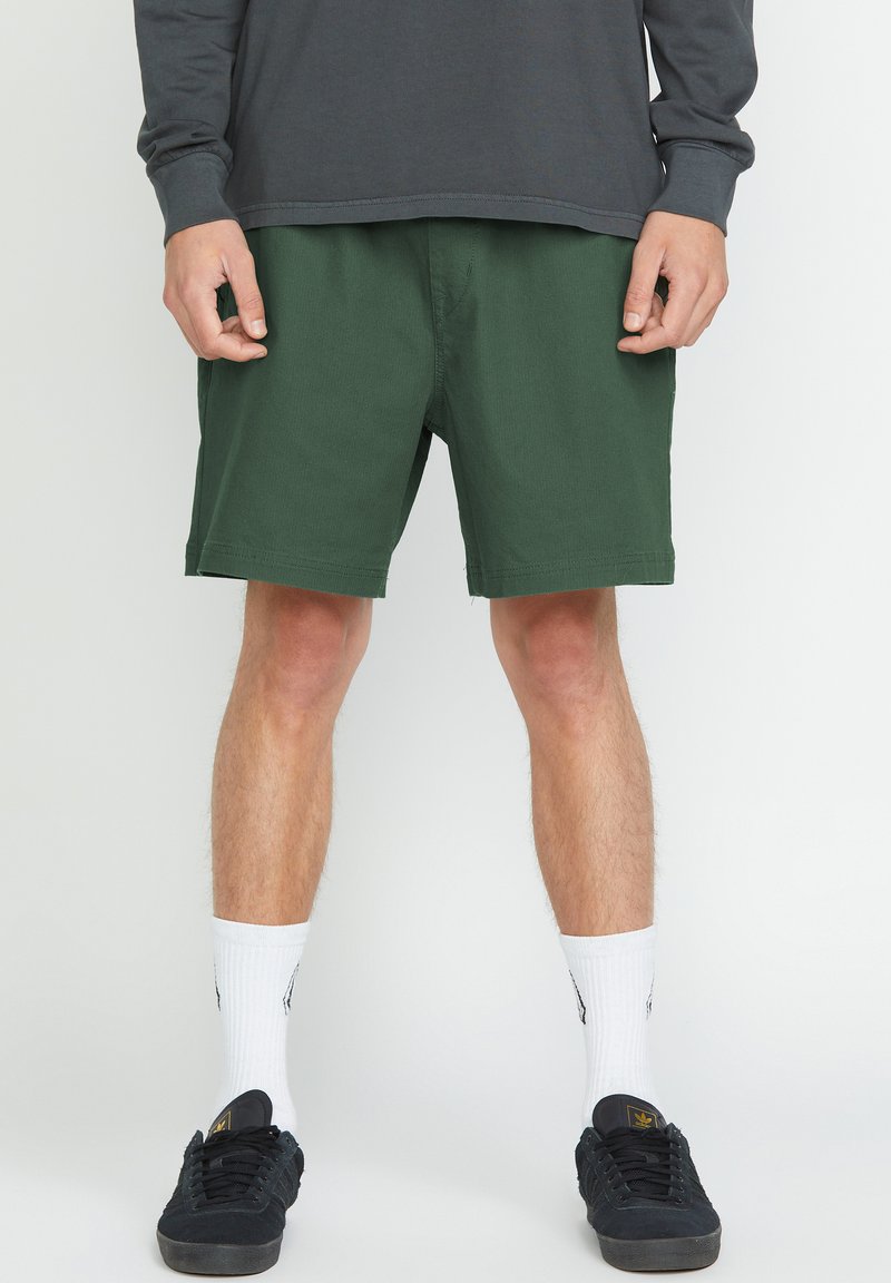 Shorts verts en coton avec une coupe décontractée. Dotés d'une taille avec une fermeture à l'avant et de poches latérales, associés à des baskets noires et des chaussettes blanches.