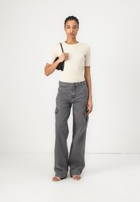 Cream short-sleeve top paired with gray cargo pants featuring pockets. Model holds a small black bag, wearing brown sandals.