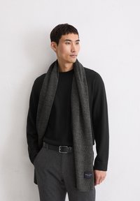 Szara, prążkowana szal w wykonany z miękkiego materiału, zarzucony na czarny sweter. Widoczna metka z brandingiem na końcu szala. W zestawie z szarymi spodniami i paskiem.