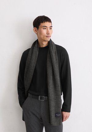 Gray ribbed scarf made of soft fabric, draped over a black sweater. Notable tag with branding on the scarf's end. Worn with gray pants and a belt.