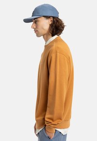 Orange sweatshirt med rund halsudskæring, ribbede manchetter og en afslappet pasform, parret med en lyseblå cap. Stoffet ser blødt og tekstureret ud.