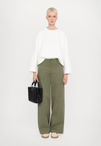 White oversized sweatshirt with embroidered logo, olive green wide-leg pants, black woven handbag, and cream-colored shoes.