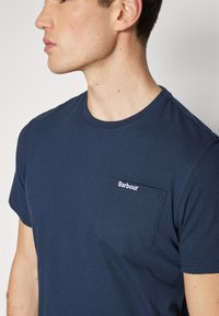 Navy blue cotton t-shirt with a crew neck and short sleeves. Features a left chest pocket with white "Barbour" logo. Smooth texture.