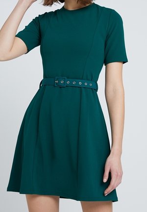 Frau trägt ein dunkelgrünes, knielanges Kleid mit kurzen Ärmeln und einem passenden Gürtel mit einer rechteckigen Schnalle.