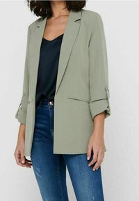 Frau in einem hellgrünen Blazer mit hochgerollten Ärmeln, schwarzem Satinoberteil und blauen Jeans vor einem schlichten Hintergrund.