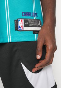 Teal basketballtrøje med hvide striber og "CHARLOTTE" i lilla. Etiket viser størrelse 48, NBA-branding og mesh-struktur. Sorte shorts med hvid swoosh.