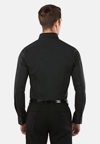 Chemise noire à manches longues avec un col structuré, texture lisse et accents violets aux poignets, portée avec un pantalon sombre et une ceinture.