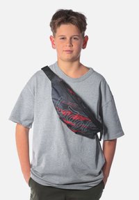 Maglietta a maniche corte grigia, borsa in vita nera e rossa con zip. La borsa è caratterizzata da un logo bianco stilizzato e una superficie in tessuto testurizzato.