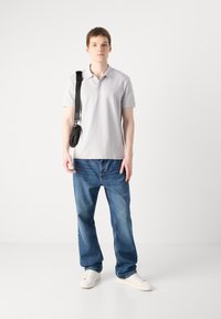 Polo gris clair à manches courtes, avec un logo discret. Associé à un jean en denim bleu et des baskets blanches. Sac noir sur l'épaule.