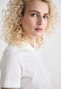Balta polo marškiniai su tekstūruotu audiniu, turintys apykaklę ir tris mygtukus. Modelis turi garbanotus šviesius plaukus, fone matoma neutrali aplinka.