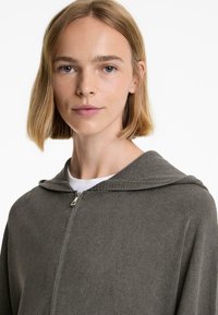 Grå zip-up hoodie med huva, med ribbad textur och en slät dragkedja. Bärs över en vit T-shirt, mot en enkel vit bakgrund.
