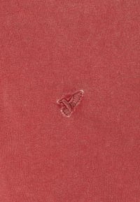 Tessuto di cotone rosso con un piccolo logo di un uccellino ricamato in rosa. Texture liscia con leggere variazioni di colore e fibra.