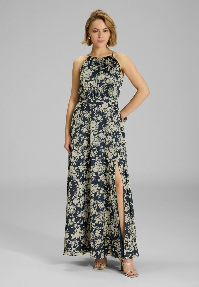 Maxi mekko tummansinisellä pohjalla ja kermavärisellä kukkakuviolla, kaula-aukko on halterneck-tyyppinen, istuva vartalonmyötäinen malli ja sivuhalkio; yhdistettynä nilkkaremmikenkien kanssa.